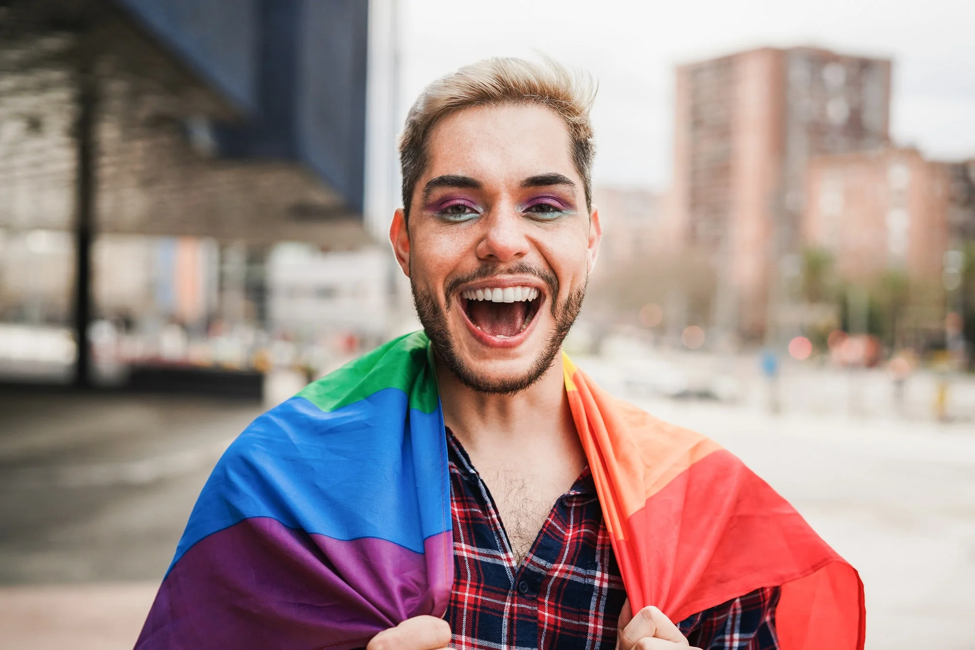 Gay man with makeup on having fun wearing lgbt rainbow flag outdoor - Focus on face